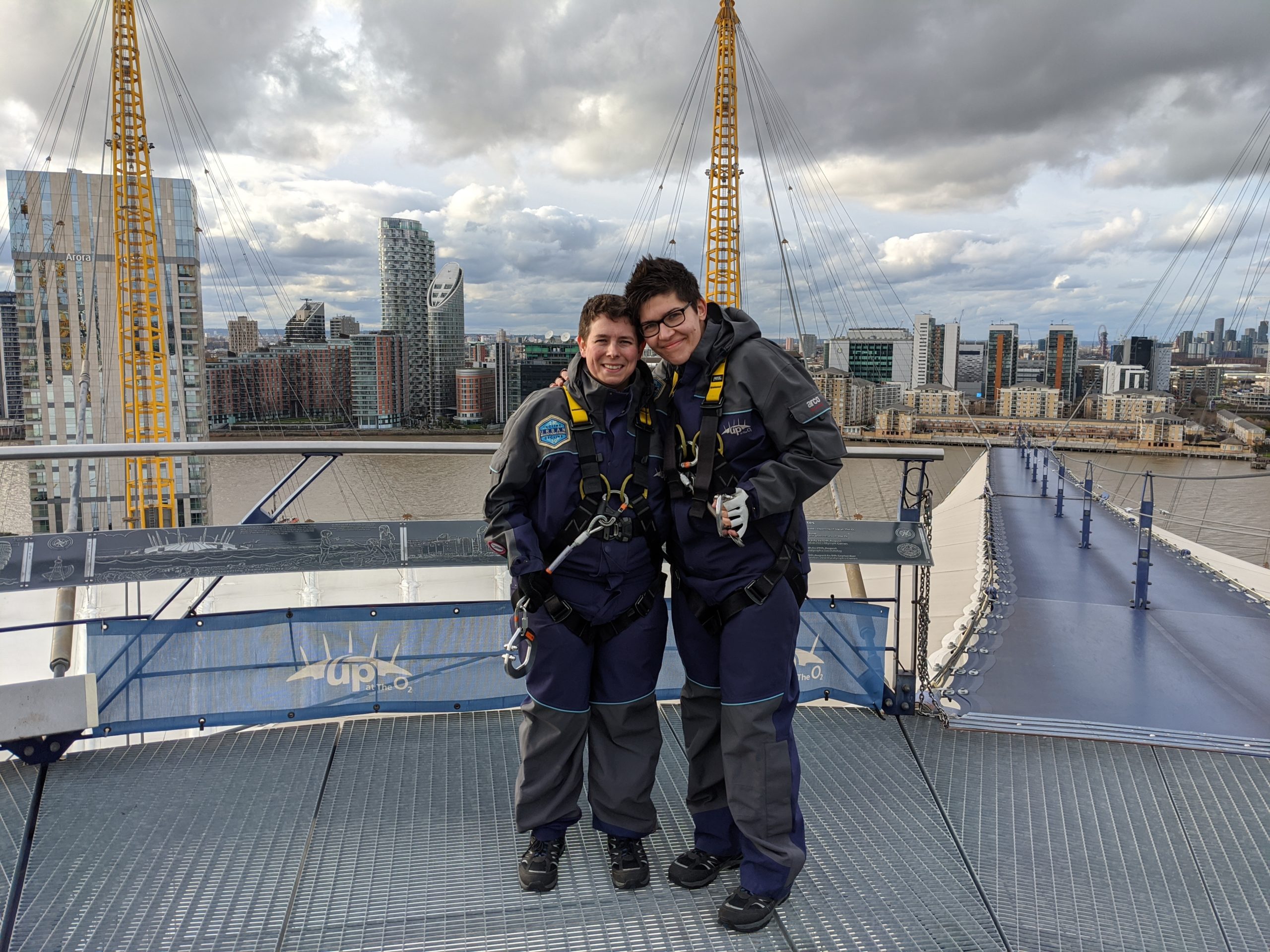 Picture of Sharon and I standing at the top of The O2 after our climb. A London Skyline sits behind us. We are dressed in climbing gear and have climbing equipment attached to us,
